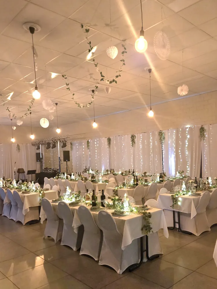 Lange tafels met witte stoelhoezen en kleine bloemstukken, onder lichtslingers en witte gordijnen.