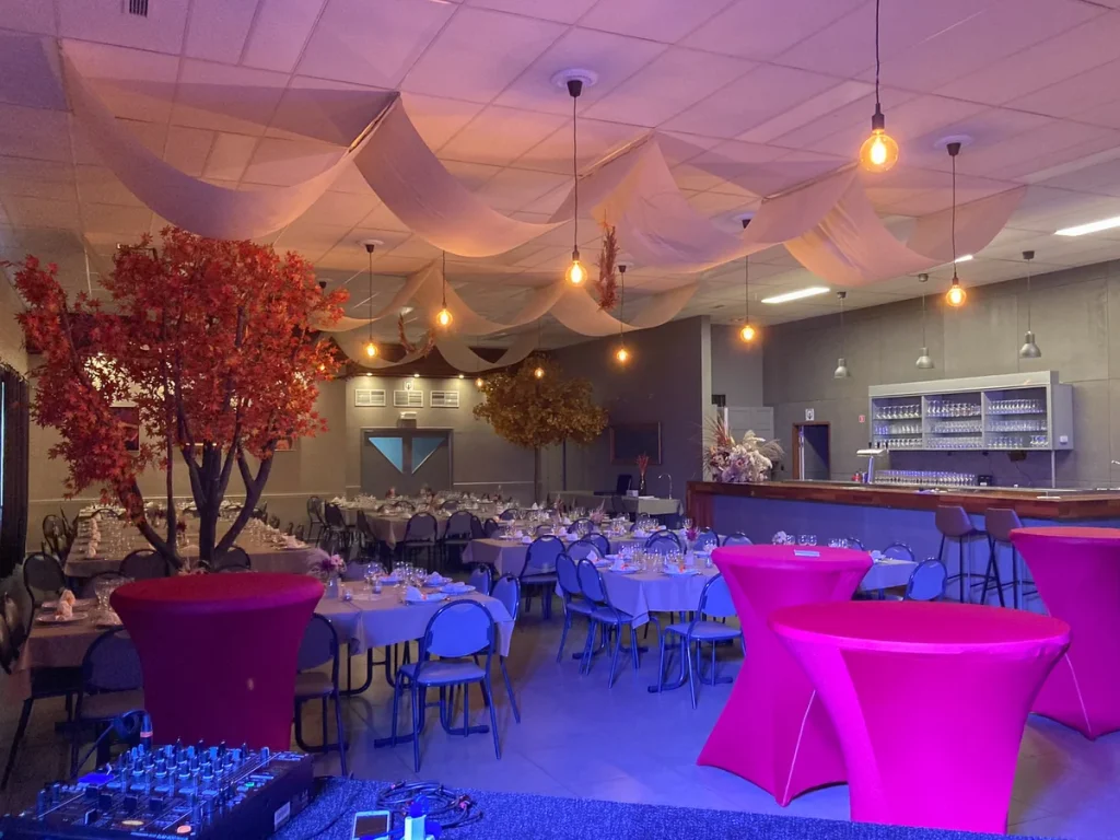 De zaal in paars en roze licht, met een DJ-booth, roze statafels, doeken aan het plafond en bloesembomen tussen de gedekte tafels.